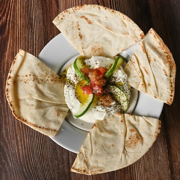 Labneh dip with olive oil, diced tomatoes, cucumbers, and pita bread on a wooden table.