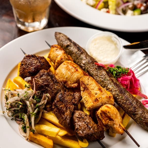 A white plate with a mixed grill of chicken and beef skewers, kofta, fries, and pickled onions.