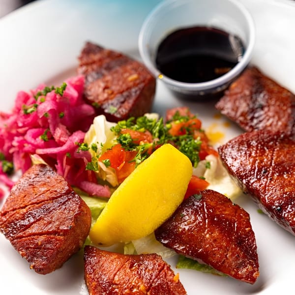 Grilled meat pieces with pickled red onions, salad, a lemon wedge, and a side of sauce.
